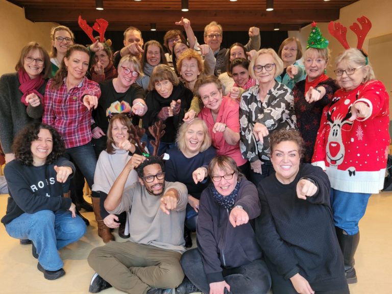 Ein Gruppenfoto der Chormitglieder. Alle zeigen auf auf den Leser/die Leserin und wünschen Frohe Weihnachten